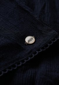 Marineblauwe textuurstof met een goudkleurige ronde knoop met het "Superdry" logo en een gegolfde randafwerking.