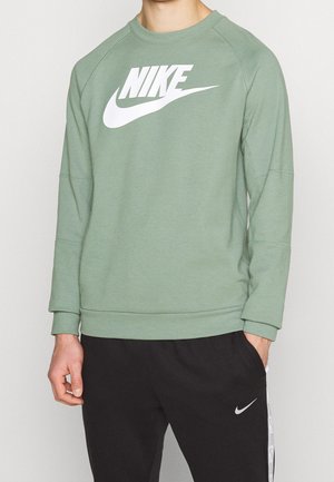 Mężczyzna ubrany w zieloną bluzę Nike z dużym białym logo oraz czarne spodnie dresowe Nike z małym białym znakiem Swoosh na lewej nodze.