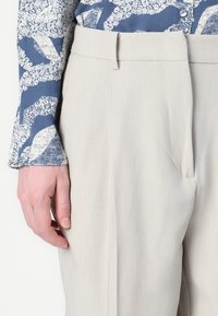 Light gray trousers with a tailored fit, featuring a flat front, belt loops, and subtle vertical stripes. A patterned blue top is partially visible.
