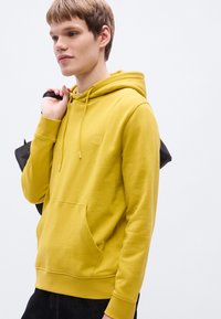 Κίτρινο φούτερ με κουκούλα από απαλό ύφασμα, με μπροστινή τσέπη, κορδόνι στην κουκούλα και μικρό λογότυπο. Το μοντέλο κρατάει μια μαύρη τσάντα.