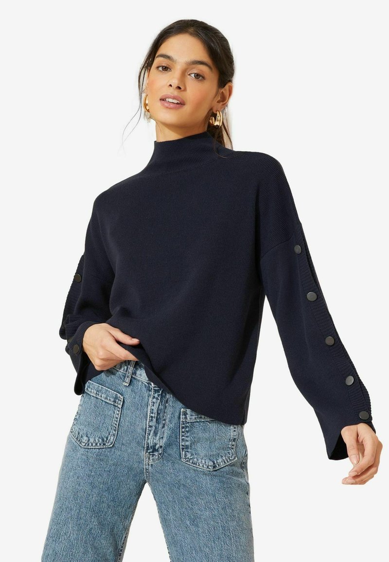 Pullover a coste blu navy con colletto alto, caratterizzato da dettagli con bottoni lungo le maniche e un fit rilassato. Abbinato a jeans in denim blu.