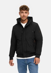 Man in zwarte winterjas met capuchon over een wit shirt en blauwe spijkerbroek, staande tegen een effen witte achtergrond.
