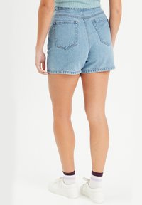 Shorts en denim bleu clair avec des bords effilés, poches arrière plates et taille haute. Portés avec des baskets blanches et des chaussettes à rayures violettes.