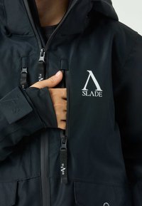 Black waterproof jacket with a high collar, front zip, adjustable cuffs, and notable logo detail on the chest. Durable fabric with a smooth texture.