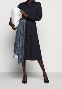 Abito camicia blu navy realizzato in tessuto morbido, con dettaglio alla vita con cintura. Abbinato a una sciarpa blu con motivo e con una borsa bianca, indossato con tacchi neri.