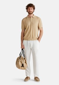 Beige knitted polo shirt with short sleeves and ribbed hem, paired with white linen pants. The model holds a textured tan duffel bag.