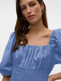 Sieviete ar gariem brūniem matiem, valkājot sudrabotas stīpu auskarus un gaiši zilu kleitu ar pūstošām piedurknēm un savilktu taisnstūrveida kakla izgriezumu.