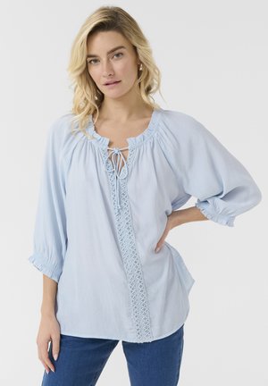 Blouse bleu clair avec un col rond, détail de cordon, et un panneau avant brodé ; présente des manches longues bouffantes avec des poignets élastiques.