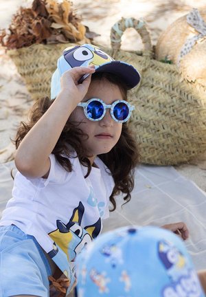 Bambino con un cappello azzurro chiaro con un design di cartone animato, occhiali da sole tondi blu e una maglietta bianca con una grafica colorata, seduto su un tappeto.