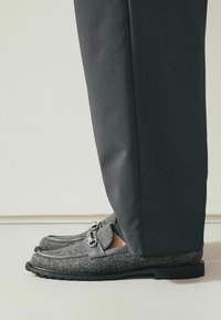 Grijze gevoerde loafers met zwarte leren accenten, voorzien van een gespsluiting. De schoen heeft een ronde neus en een grove textuur. Gepaard met donkere broeken.