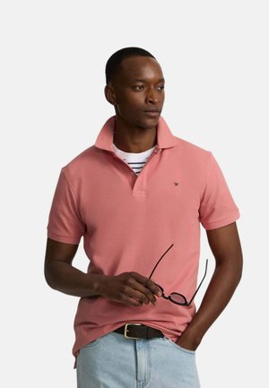 Hombre con una camisa polo rosa sobre una camiseta de rayas, vaqueros azul claro con un cinturón negro, sosteniendo unas gafas con montura negra mirando hacia un lado.