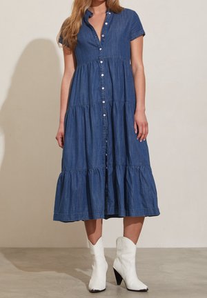 Kobieta ubrana w niebieską sukienkę na guziki o długości do połowy ciała i krótkim rękawie, w parze z białymi botkami na obcasie sięgającymi połowy łydki, stojąca na gładkiej podłodze.