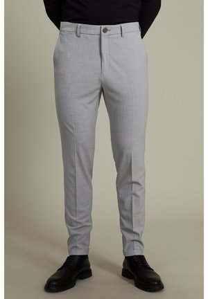 Schmal geschnittene hellgraue Anzughose, getragen mit schwarzen Anzugschuhen und einem schwarzen Oberteil, stehend vor einem schlichten beigen Hintergrund.