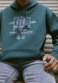 Tmavě zelená mikina s kapucí s potiskem na přední straně "Digital Deviance." Má šňůrky na stažení, přední kapsu a žebrované manžety.