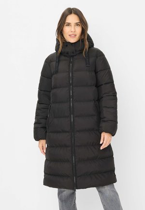 Frau trägt einen langen schwarzen Steppmantel mit hohem Kragen, Frontreißverschluss und Seitentaschen, steht vor einem schlichten hellen Hintergrund.