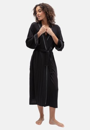 Femme aux cheveux bouclés portant une longue robe noire avec des bordures blanches, pieds nus, tenant le col tout en regardant vers le bas sur le côté.