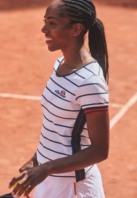 Camisa de tenis blanca a rayas con cuello redondeado, logo de marca y mangas cortas con acentos en rojo y azul. Fondo de cancha de tenis.