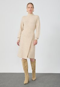Abito lavorato a maglia beige con colletto alto, maniche lunghe e design a incrocio diagonale. Abbinato a stivali in camoscio beige al ginocchio.