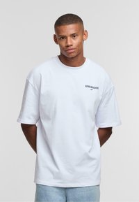 White oversized t-shirt made of cotton. Features short sleeves and a small printed text "ASPIRE AND ACHIEVE" in dark blue on the chest.