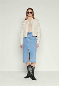 Mulher vestindo uma jaqueta bomber bege, blusa bege, saia midi em denim azul com fenda frontal, botas de cowboy pretas bordadas e óculos de sol redondos.
