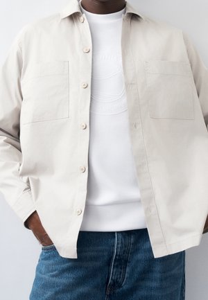 Hombre con camisa beige clara abotonada sobre sudadera blanca y vaqueros azules con las manos en los bolsillos, recortado a la altura del cuello y la cintura.