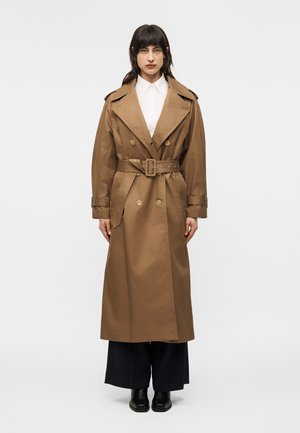 Donna in piedi che indossa un lungo trench marrone con cintura sopra una camicia bianca e pantaloni neri a gamba larga con scarpe nere, su uno sfondo chiaro e uniforme.
