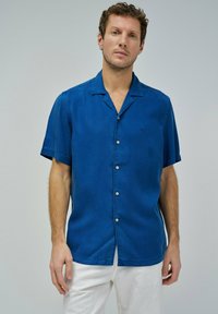 Camisa de manga corta, con cuello, en tejido azul oscuro con una textura suave, cierre de botones al frente y detalle sutil del logo. Combinada con pantalones blancos.
