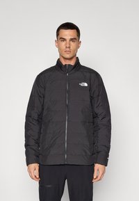Jachetă neagră cu umplutură, cu guler înalt, închidere completă cu fermoar și logo pe pieptul stâng. Material texturat cu modele cusute orizontal.