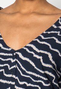 Primo piano del collo e della parte superiore del torace di una persona che indossa una camicia blu navy con strisce ondulate bianche e azzurre chiaro e bottoni neri.