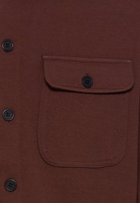 Bruine button-up shirt met een zak, vervaardigd van een gestructureerde stof en zwarte knopen. De zak is vierkant van vorm en heeft een klep sluiting.