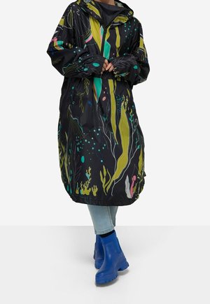 Person iført lyseblå jeans, en sort regnfrakke med grønne og farverige plante-mønstre samt klare blå ankelregnstøvler.