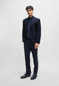 Abito blu marina con taglio slim, giacca monopetto e pantaloni abbinati, indossato con una camicia scura e una cravatta, completato da scarpe in pelle nera.