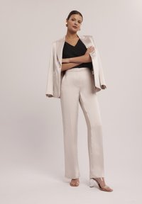 Jasnobeżowa satynowa marynarka i dopasowane spodnie zestawione z czarnym topem w kształcie litery V. Przezroczyste sandały na obcasie dopełniają całość.