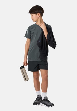 Chłopiec w szarej sportowej koszulce i spodenkach trzyma czarną kurtkę zarzuconą na ramię, a w drugiej ręce butelkę wody z napisem „Swedemount”.