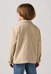Beige Canvasjacke mit Kragen, Knopfärmeln und lockerer Passform, getragen über blauen Jeans. Sichtbare Nähte auf der Rückseite.