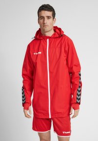 Rød letvægtsjakke med hætte, der har en frontlynlås, elastiske manchetter og sorte chevrondesign på ærmerne. Båret over matchende shorts.