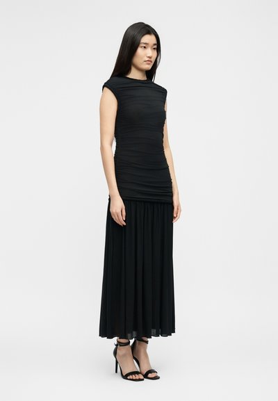 Γυναίκα που στέκεται φορώντας μαύρο τοπ χωρίς μανίκια με πιέτες, μακριά πλισέ μαύρη φούστα και μαύρα πέδιλα με λουράκι στο αστράγαλο και ψηλά τακούνια πάνω σε απλό φόντο.