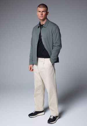 Junger Mann steht mit den Händen in den Taschen, trägt graue Jacke, schwarzes Hemd, cremefarbige Hose und schwarze Sneakers vor einem einfarbigen grauen Hintergrund.