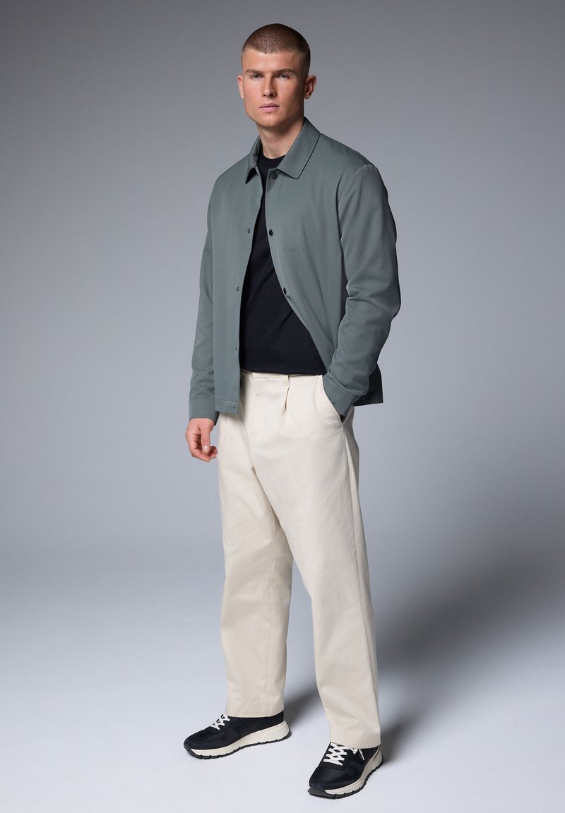 Joven de pie con las manos en los bolsillos, vestido con chaqueta gris, camiseta negra, pantalones color crema y zapatillas negras sobre un fondo gris liso.