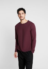 Langärmliges Rundhals-T-Shirt aus burgunderfarbigem Stoff. Verfügt über eine lockere Passform und eine glatte Textur. Kombiniert mit schwarzen Hosen.
