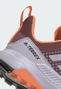 Zapato de senderismo Terrex con una parte superior de malla color marrón burdeos, acentos naranjas, superposiciones sintéticas grises y una suela exterior texturada y duradera con tacos de tracción negros.