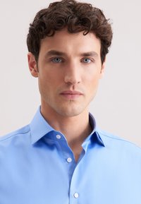 Camicia azzurro chiaro con colletto a sbuffo, realizzata in tessuto morbido. Presenta bottoni bianchi e un design aderente.