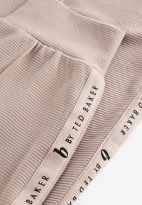 Raukšlėtas smėlio spalvos audinys su juodu „by Ted Baker“ logotipu palei kraštą. Dizainas pasižymi minkšta tekstūra ir subtiliais klostuotės akcentais.