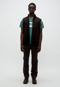 Μαύρο γιλέκο από φλίς πάνω από μια πετρόλ μπλούζα με λευκό λογότυπο, συνδυασμένο με μαύρο παντελόνι και μαύρα παπούτσια. Τα γυαλιά ηλίου ολοκληρώνουν το outfit.