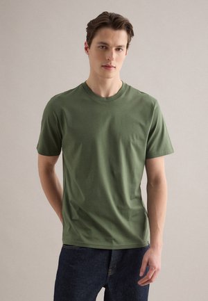 Jonge man met een olijfgroene T-shirt met korte mouwen en een donkere spijkerbroek, staand tegen een effen lichte achtergrond.