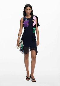 Marineblaues Kleid mit asymmetrischem Saum, floralem Muster in Pink und Grün, ärmelloses Design, kombiniert mit einer kleinen Tasche und High Heels.