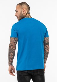 Trumpas rankovėmis mėlynas marškinėlis, medvilnės medžiaga, vienspalvis dizainas, apvalus iškirpimas, vaizdas iš nugaros su matomomis tatuiruotėmis ant rankų.