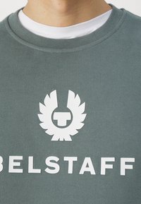 Primer plano de una persona que lleva una sudadera verde con un logo y letras blancas de Belstaff en el pecho sobre una camisa blanca.