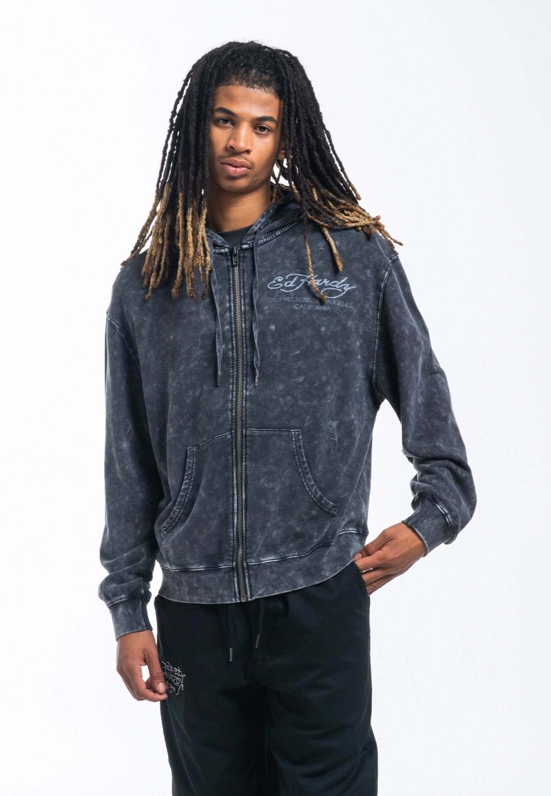 Sudadera con cremallera de lavado ácido negro con cordón, bolsillos frontales y logo bordado. Textura suave con puños y dobladillo acanalados.