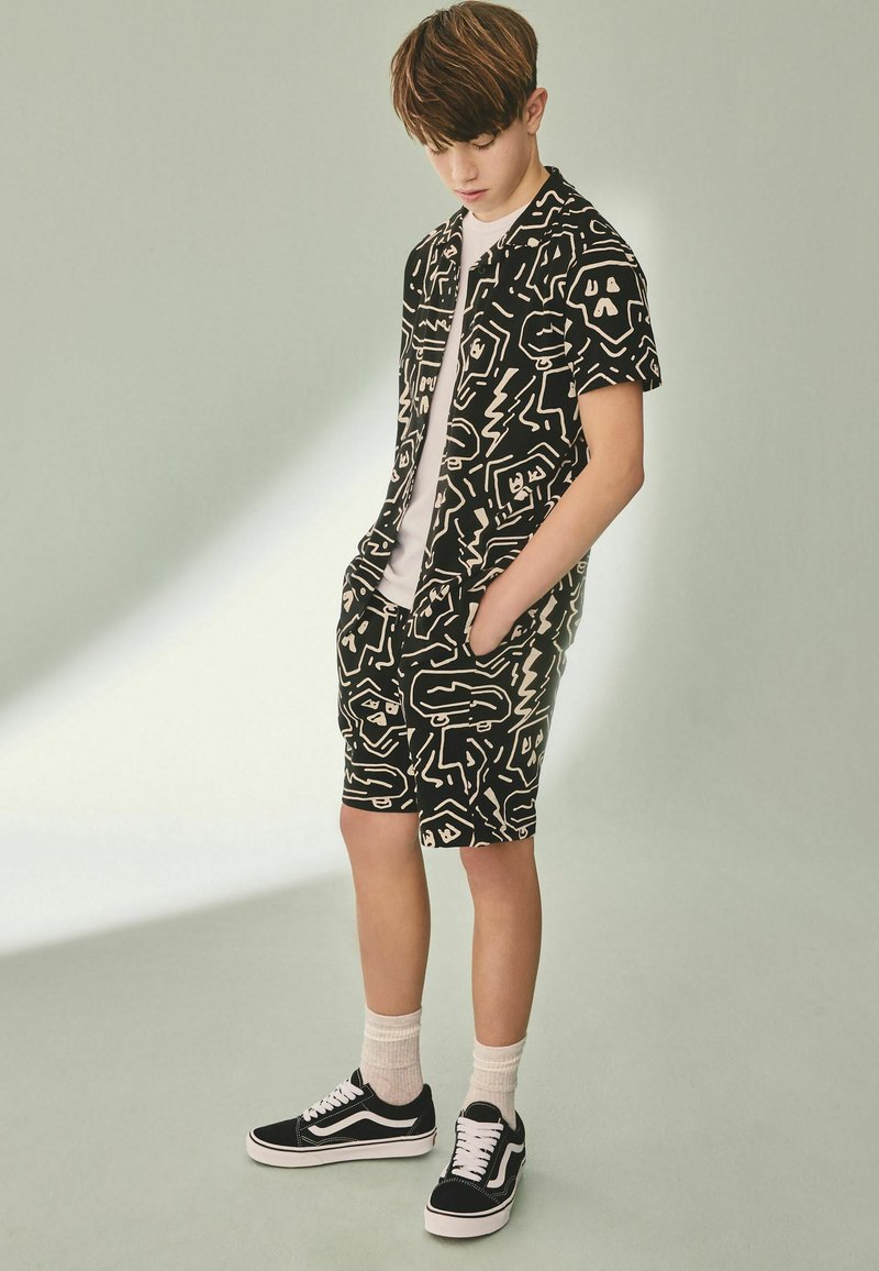 Teen boy wearing black and white patterned short-sleeve shirt and matching shorts, white socks, and black sneakers, looking downward with hands in pockets.
